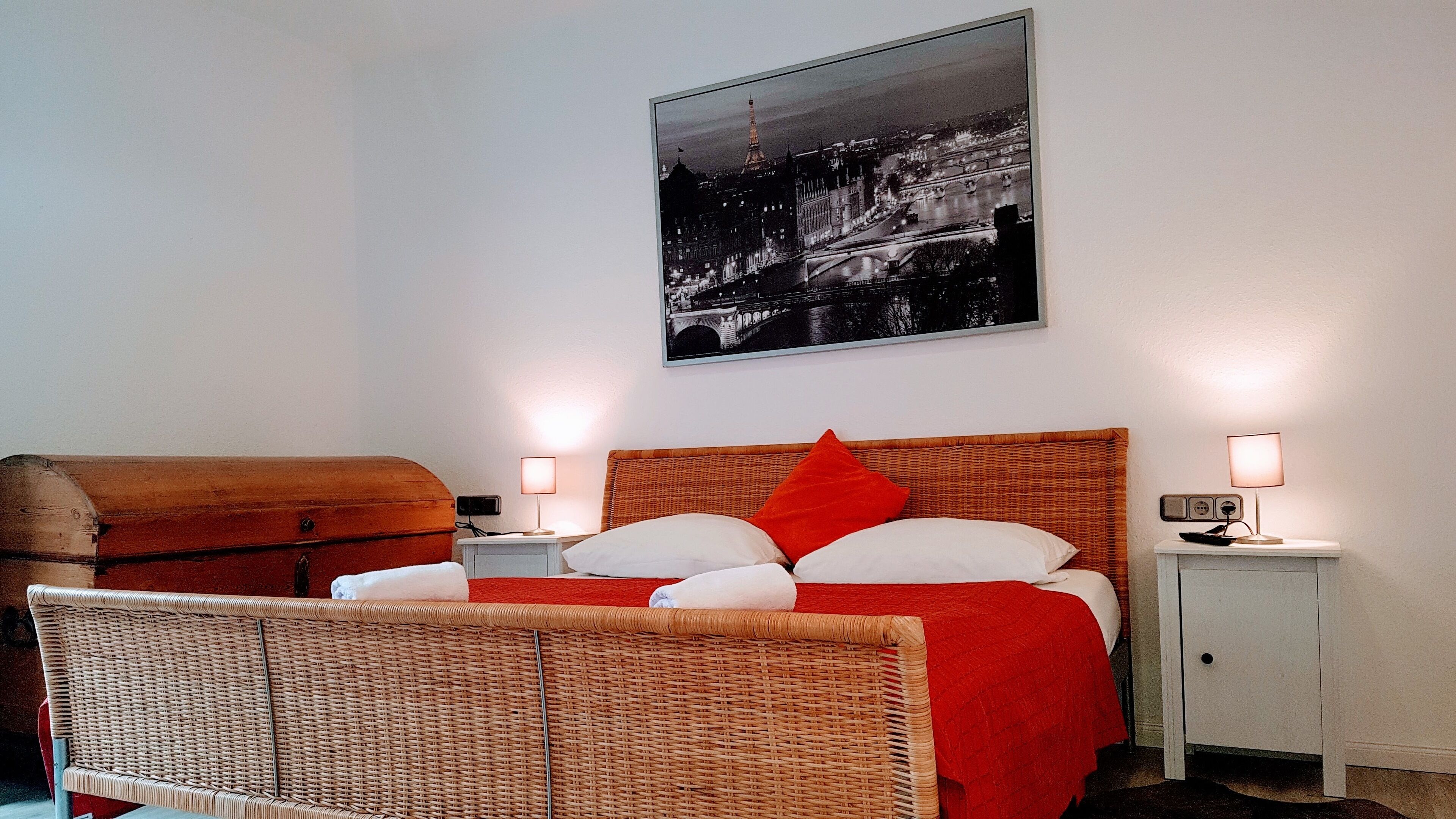 Habitación de hotel con cama, una silla de mimbre, mesita de noche, lámpara y un cuadro enmarcado con vista a la ciudad.