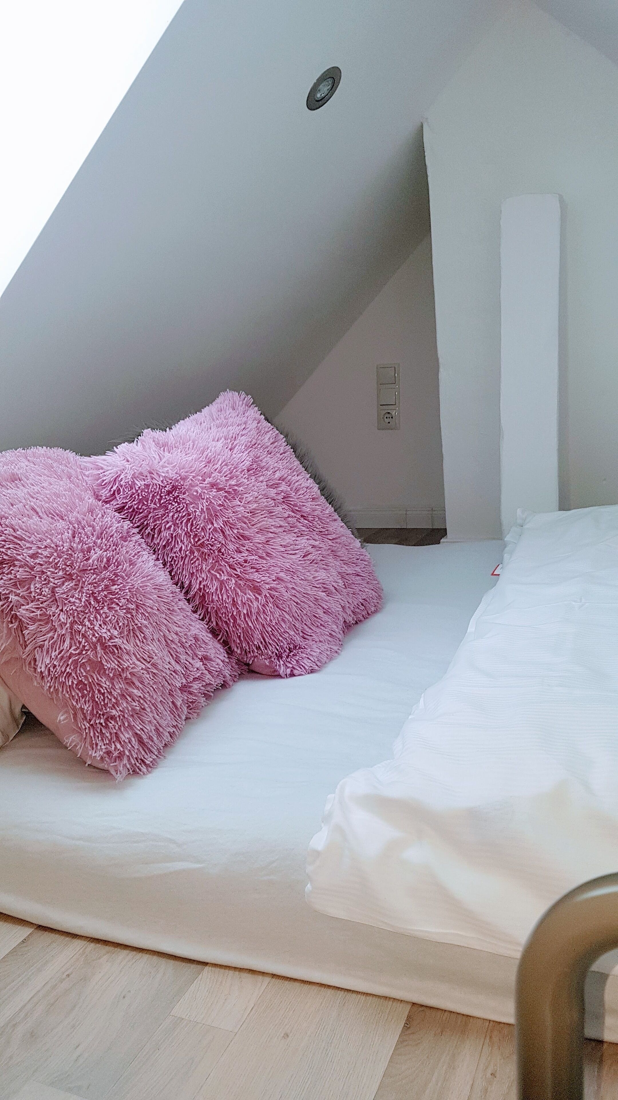 A bedroom with a sloped ceiling, a bed with white bedding, and pink fluffy pillows.