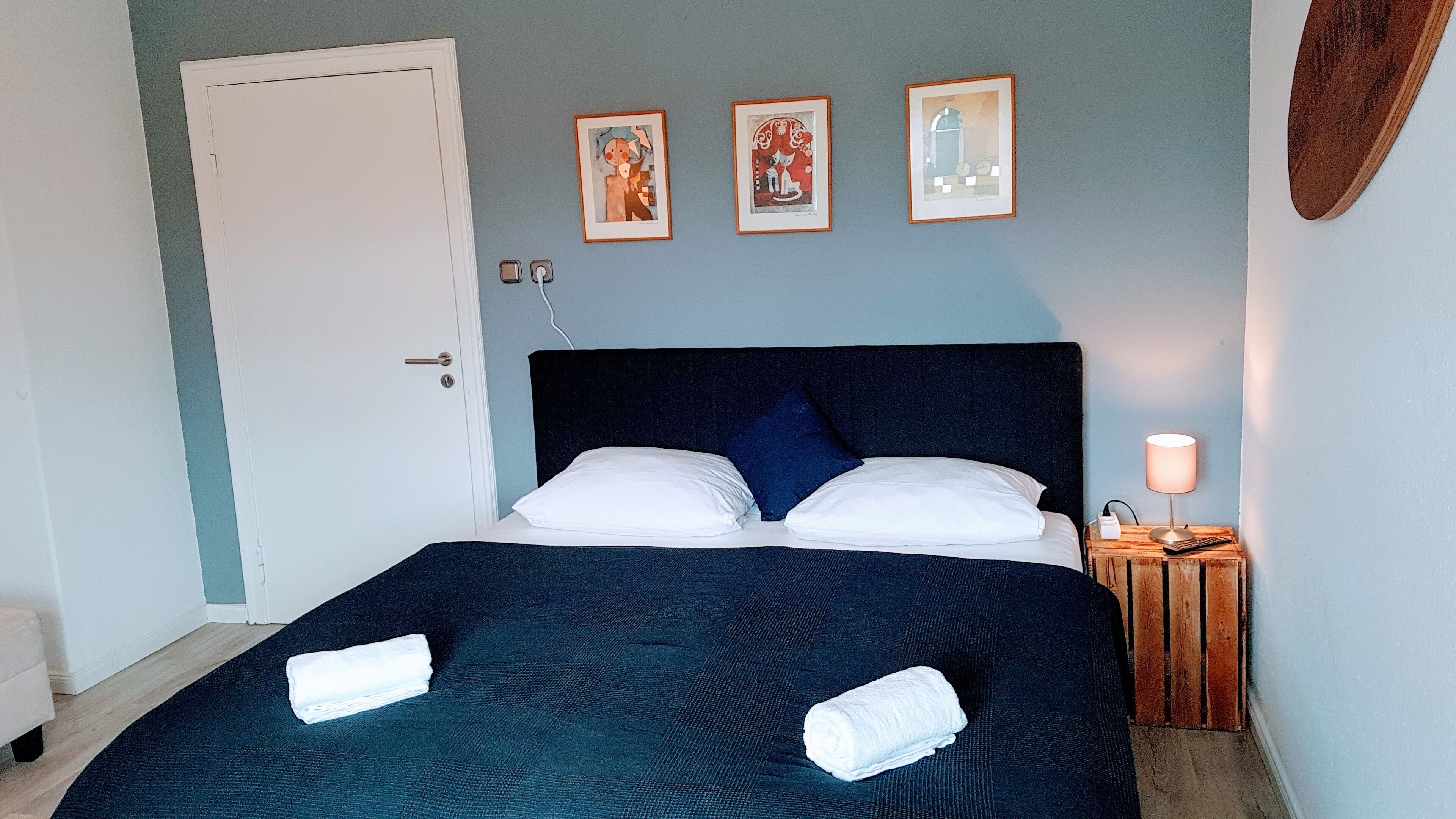 A neatly made bed with two white pillows and two folded towels, a bedside table with a lamp, and three framed artworks on the wall.