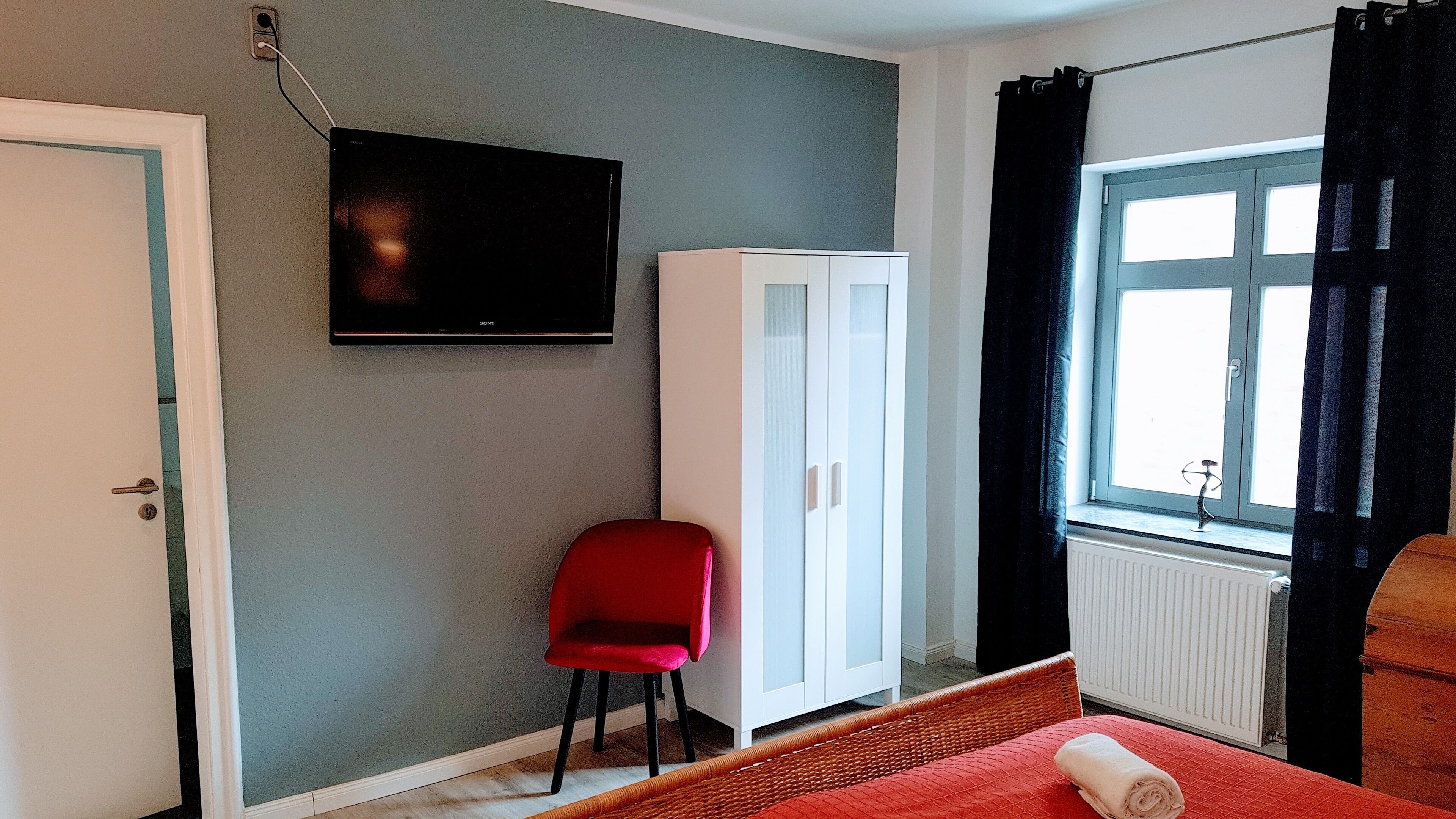 A room with a television mounted on the wall, a white wardrobe, a red chair, a window with black curtains, and a bed with a rolled towel.