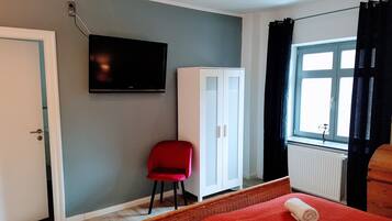 A room with a television mounted on the wall, a white wardrobe, a red chair, a window with black curtains, and a bed with a rolled towel.