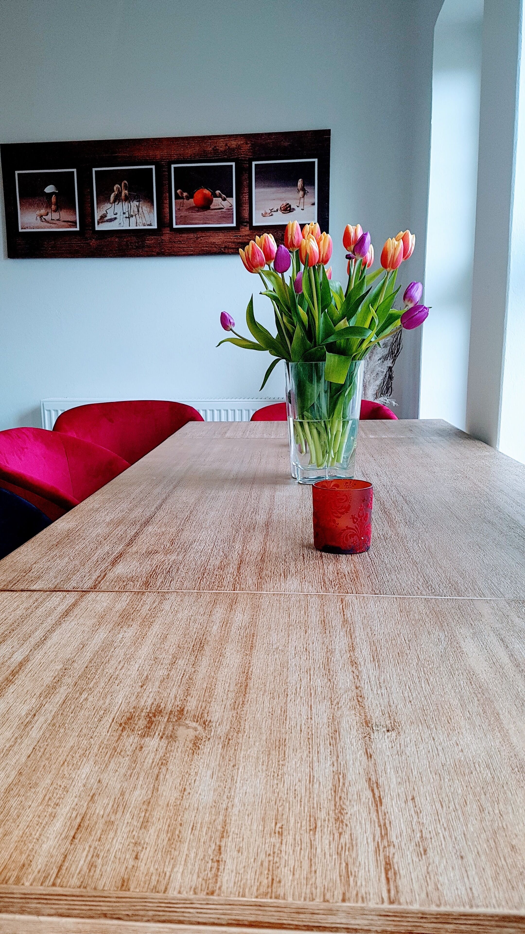 A dining table with a vase of tulips, a red candle, and a framed picture on the wall.