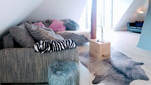 Ein gemütliches Schlafzimmer im Loft-Stil mit schrägverdeckter Decke, einem großen Fenster, einer Couch mit verschiedenen Kissen, einem kleinen Holztisch und einem einzigartigen Teppich mit Tiermotiv.