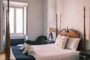 A hotel room with a four-poster bed, a desk with a lamp, a chair, and a window with blinds.