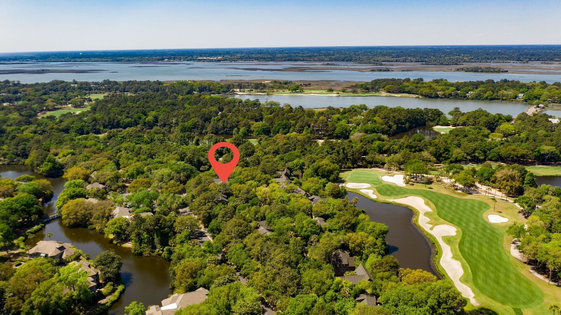Vista aérea de um campo de golfe com um marcador de localização vermelho, rodeado por florestas densas e corpos d'água.
