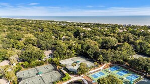 Vue aérienne d’un quartier résidentiel avec plusieurs courts de tennis, entourés d’une végétation luxuriante, et une plage en arrière-plan.