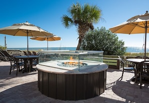 Relax in a scenic outdoor seating area with a central fire feature, tables, and chairs, surrounded by shade and a beautiful view.