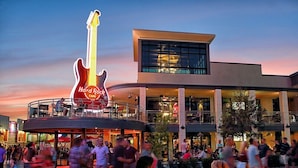 Un Hard Rock Cafe con un gran letrero de guitarra, mesas al aire libre y un edificio moderno con ventanales amplios.