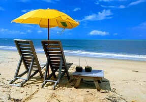 Dos sillones de madera y una mesita baja con dos bebidas de coco sobre una playa arenosa con un paraguas amarillo.