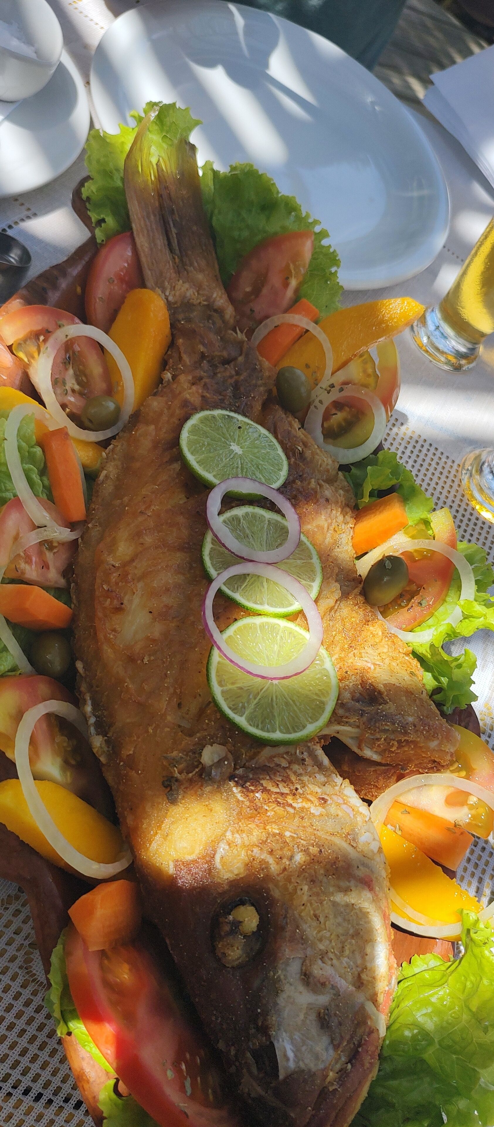 Un pescado frito entero con cebollas en rodajas, tomates y lima en un plato blanco.