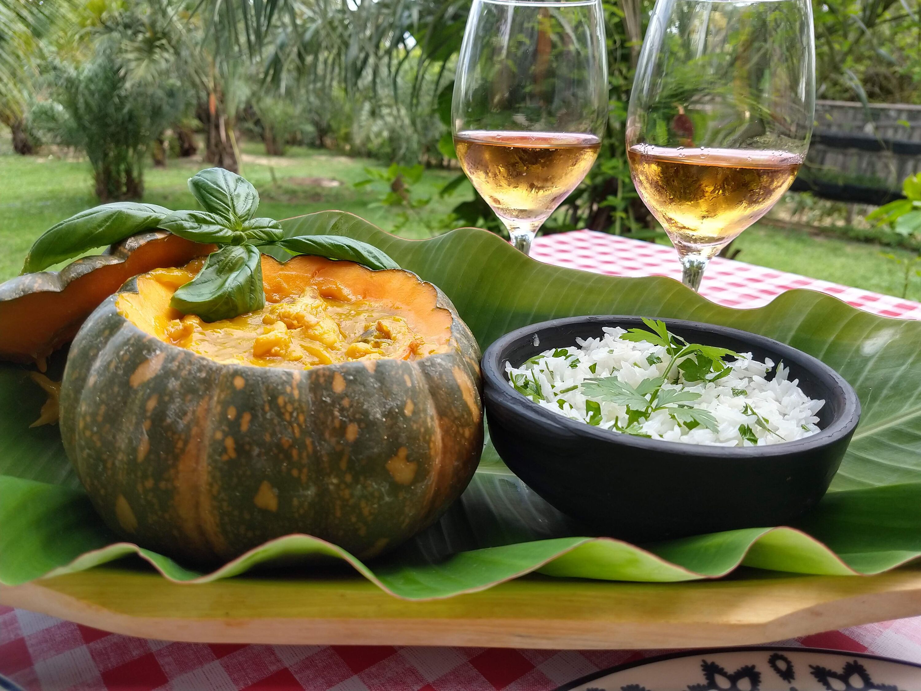 Una mesa con mantel a cuadros, un cuenco de arroz blanco adornado con hierbas, y una calabaza tallada con relleno. También se ven dos copas de vino blanco.