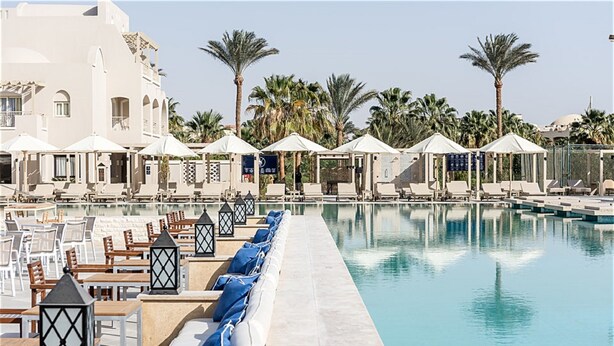 A poolside area with lounge chairs and umbrellas, surrounded by palm trees and a building with a white facade. - Jaz Makadi Gardens - TUI BLUE Makadi Gardens - Adults Friendly 16 Years Plus (Makadi Bay)