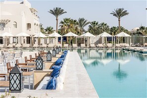 A poolside area with lounge chairs and umbrellas, surrounded by palm trees and a building with a white facade. - Jaz Makadi Gardens - TUI BLUE Makadi Gardens - Adults Friendly 16 Years Plus (Makadi Bay)