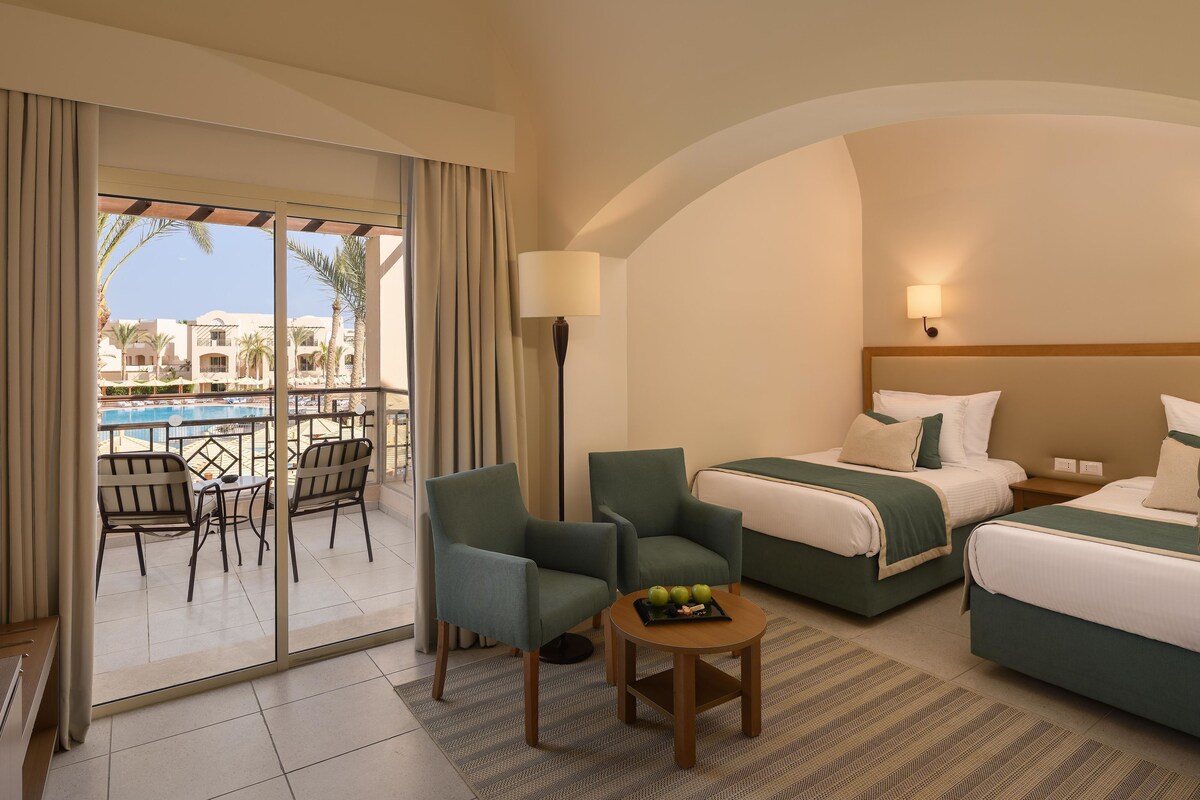A hotel room with two beds, a balcony with a pool view, a dining table with chairs, and a lamp.