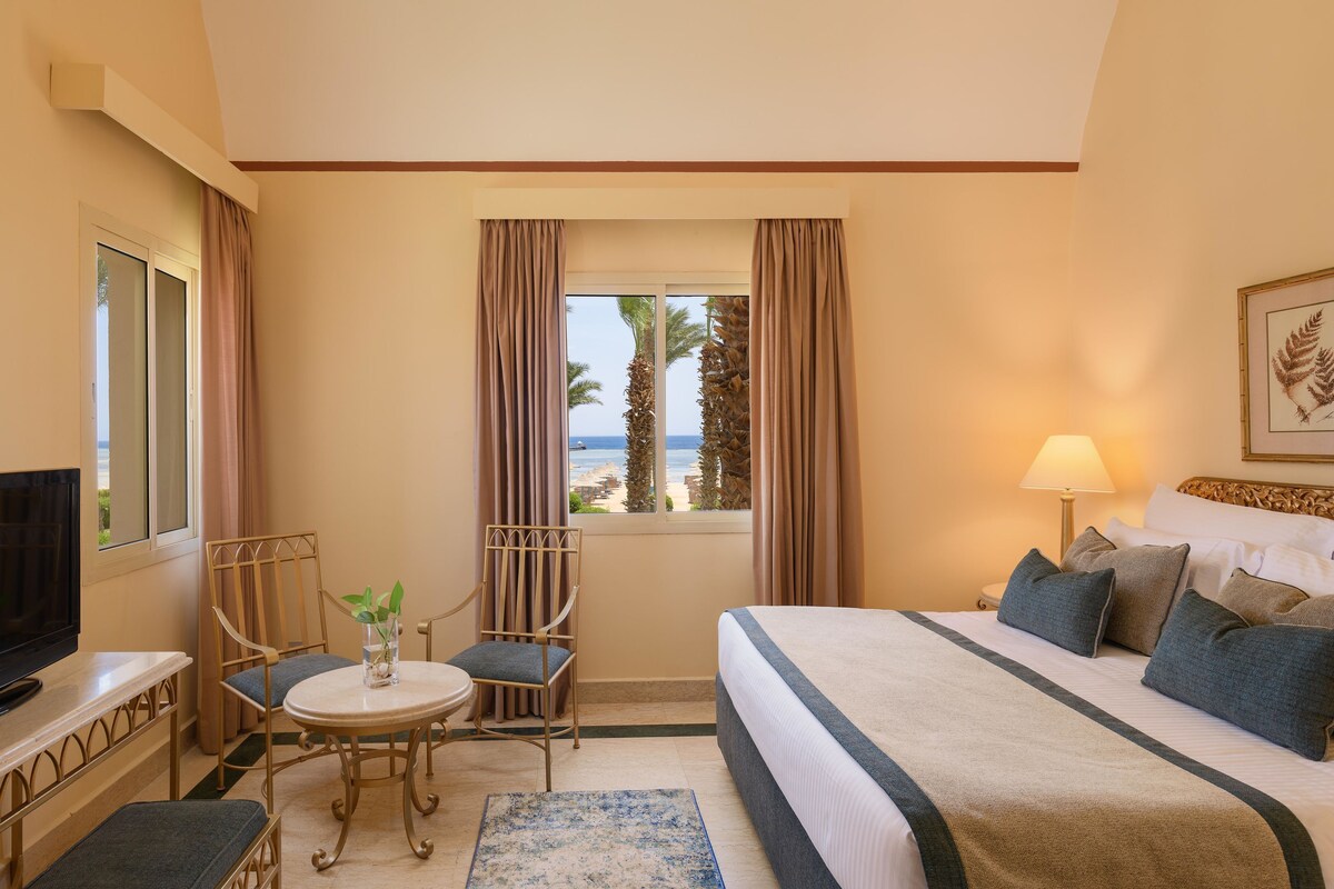 A hotel room with a bed, a television, a dining table with chairs, a window with curtains, and a lamp.