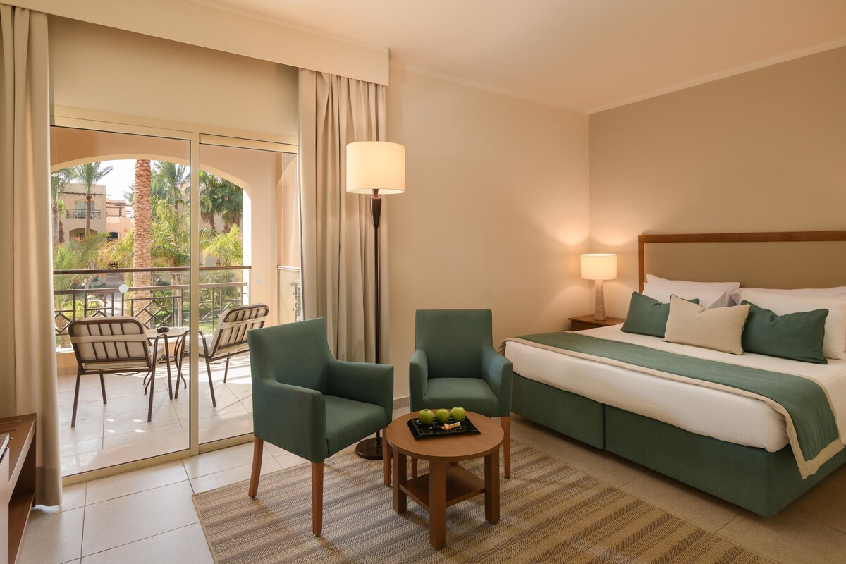 A hotel room with a bed, two chairs, a small table with fruit, a lamp, and a view through a glass door.