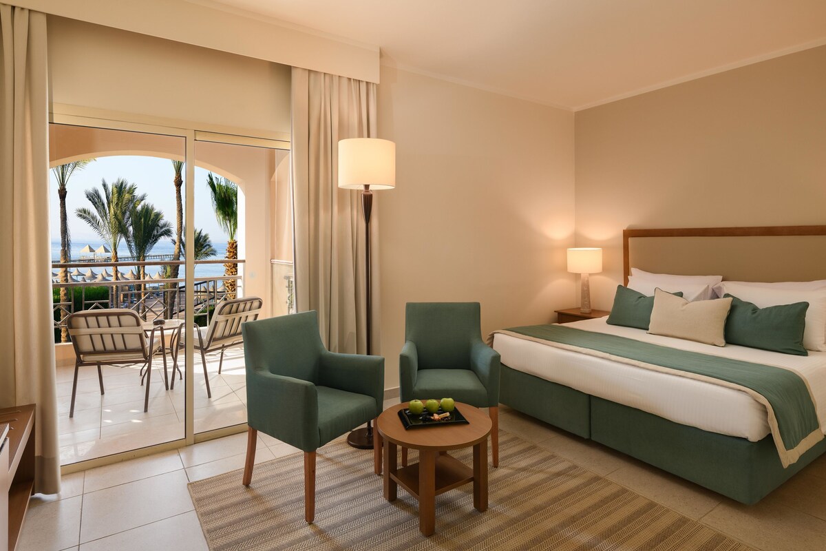 A hotel room with a bed, two chairs, a small table with fruit, a lamp, and a view of the ocean through a glass door.