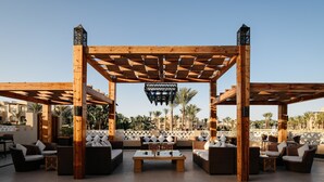 Una pérgola de madera con asientos y una mesa, palmeras al fondo y cielo despejado.