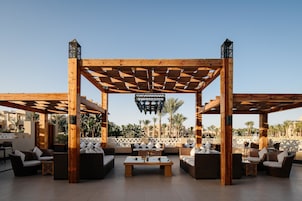 Uma pérgola de madeira com assentos e uma mesa, palmeiras ao fundo e céu claro.