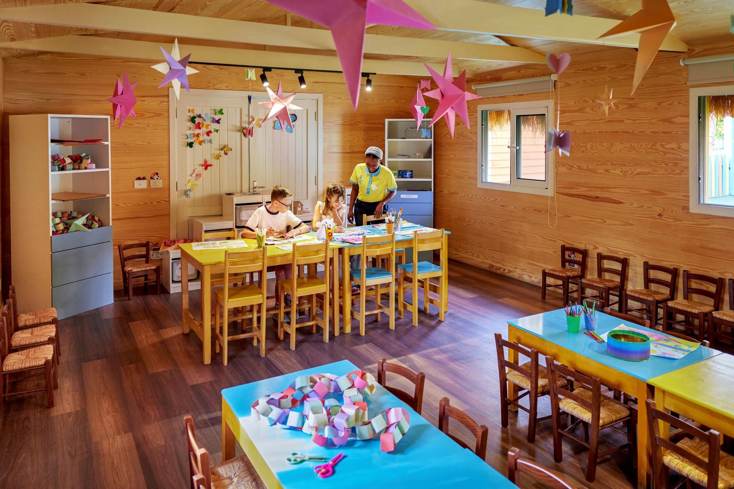 A room with wooden walls, a central table with chairs, and shelves with various items.