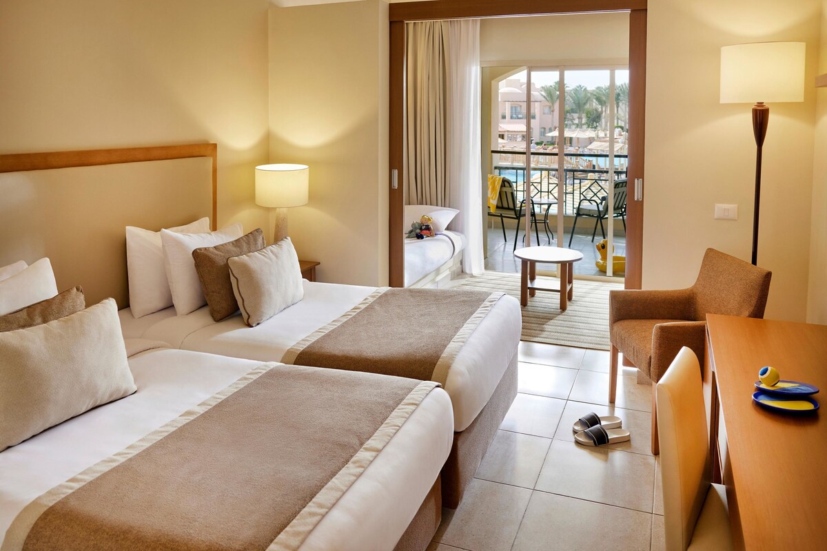 A hotel room with two beds, a balcony, a table, and a chair.