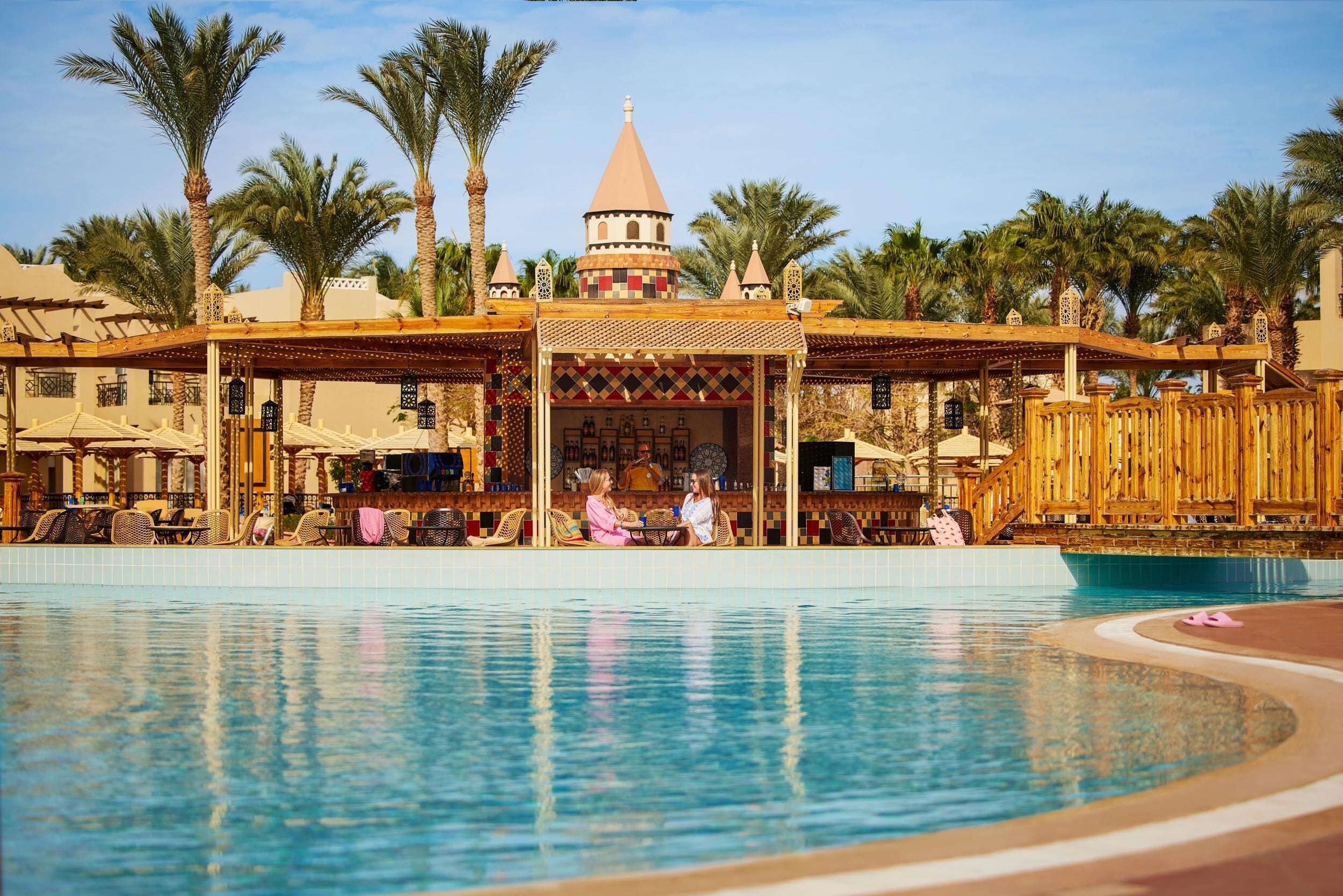 A pool area with a covered seating area, palm trees, and a distinctive building with a conical roof.