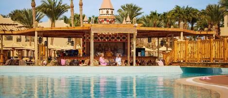 A pool area with a covered seating area, palm trees, and a distinctive building with a conical roof.