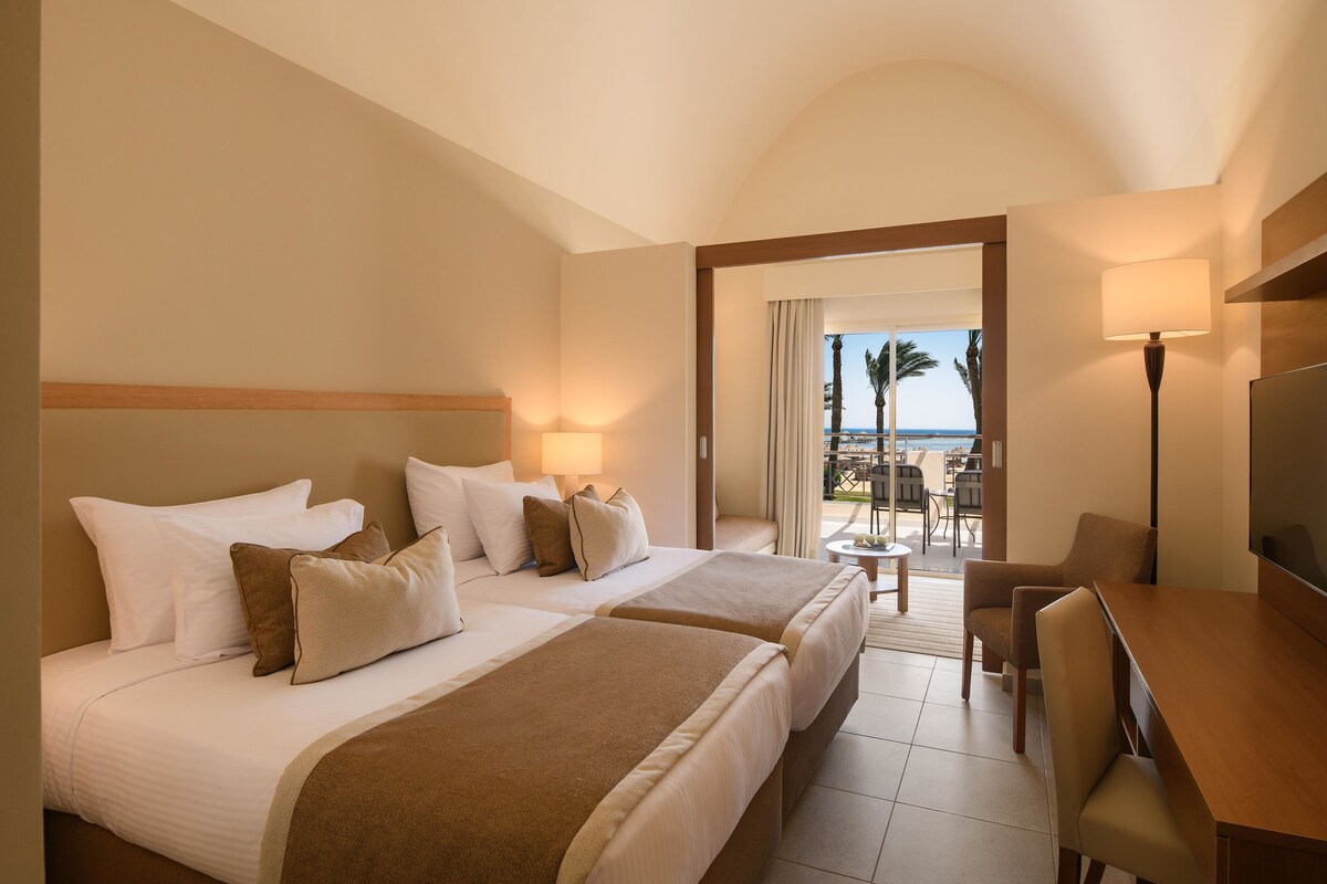 A hotel room with two beds, a desk, a chair, and a TV. There is a balcony with a view of palm trees and the sea.