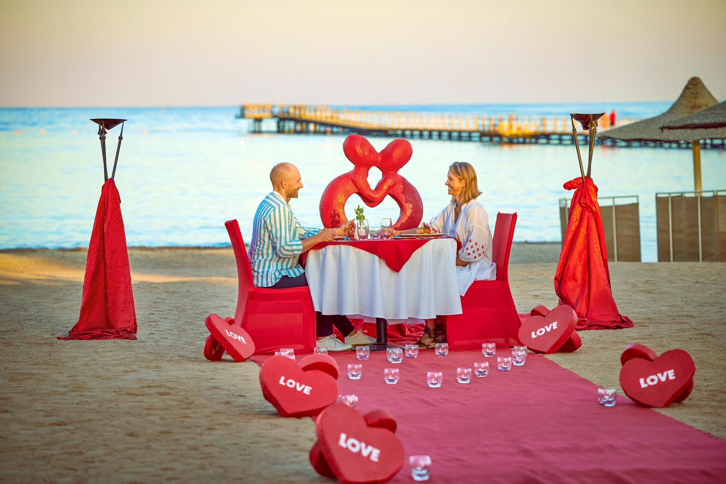 A couple dining at a beachside table with a view of the ocean and pier.