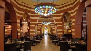 Un comedor amplio y bien iluminado con paredes a rayas y una lámpara central. Las mesas están ordenadas con sillas, y el suelo está cubierto de baldosas.