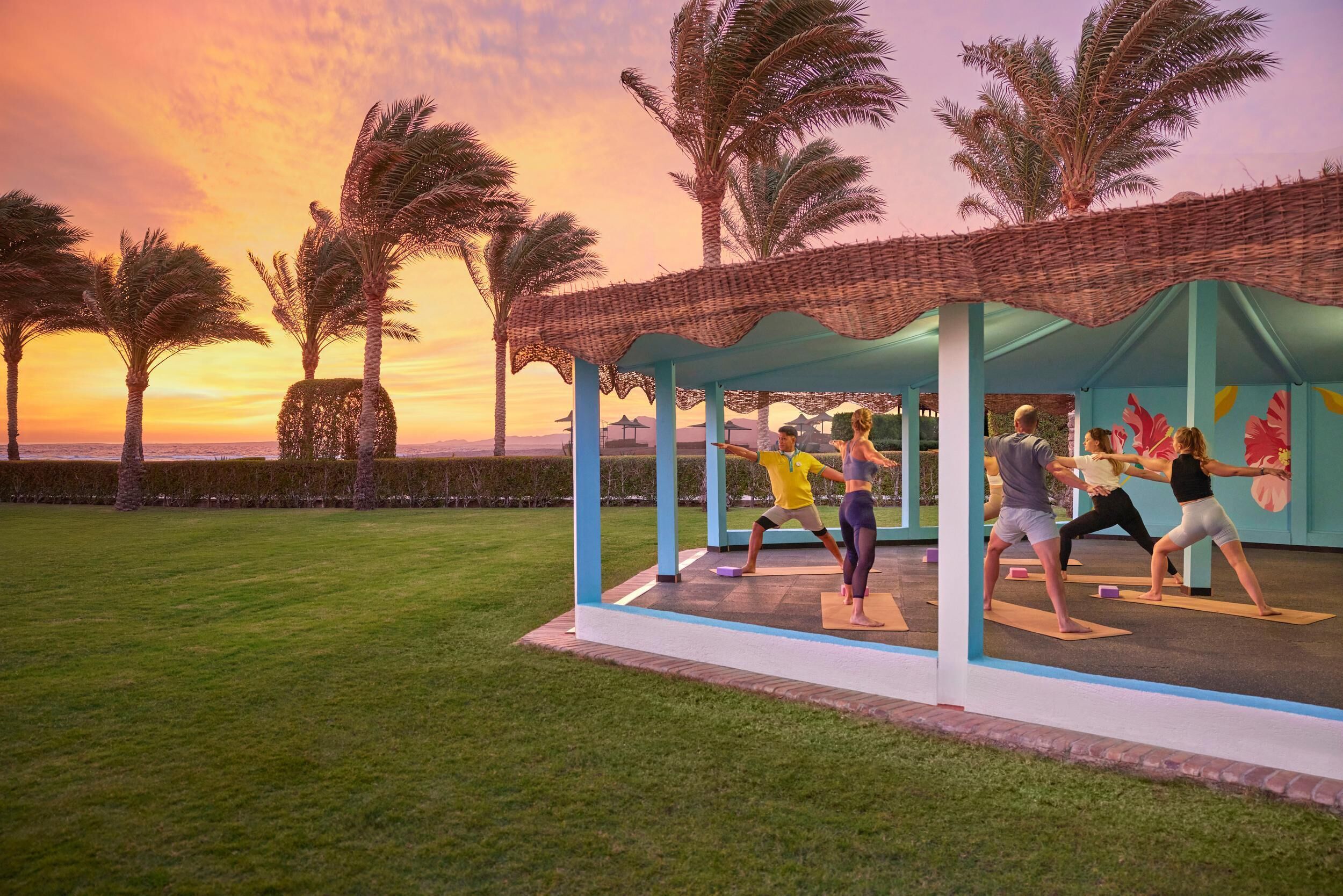 Espaço coberto ao ar livre com colchonetes de exercícios, palmeiras e vista para o pôr do sol.