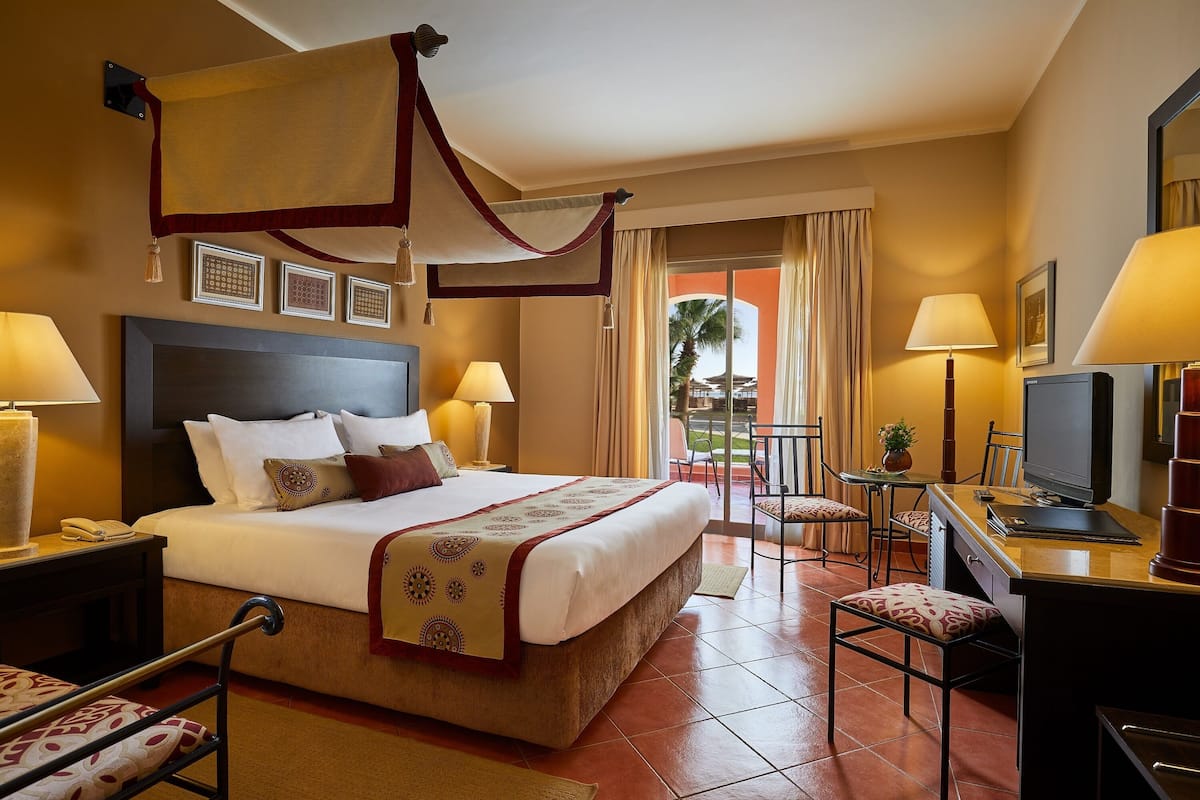 A hotel room with a large bed, two bedside tables with lamps, a desk with a computer, and a view of the outdoors.