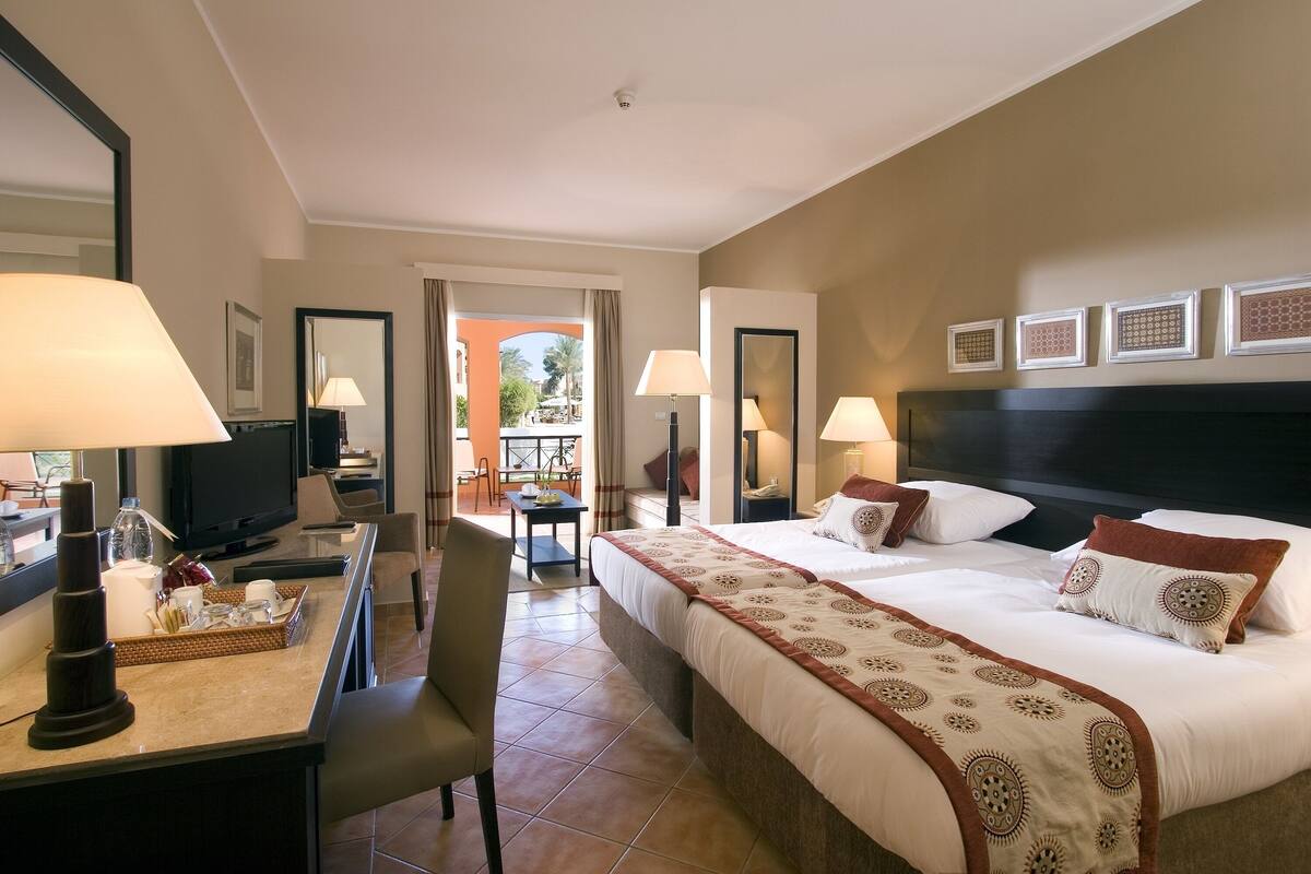 A hotel room with a bed, bedside tables, a desk with a lamp, a TV, and a balcony with a view.