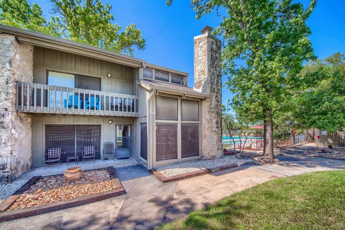 A two-story house with a stone chimney, a balcony, and a pool in the background.