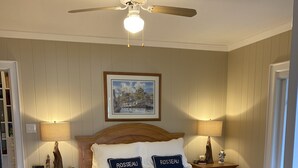 Une chambre avec un lit en bois, deux lampes de chevet, un ventilateur de plafond, un tableau encadré au mur et une table de chevet en bois avec une radio.