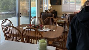 Un coin repas avec une table dressée pour quatre personnes, entourée de chaises en bois. Une bougie verte est posée sur la table, et une fenêtre avec des stores se trouve à gauche.