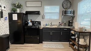 Cocina compacta con un refrigerador, estufa y microondas negros. Hay dos ventanas con persianas, un reloj en la pared y una pequeña repisa con vajilla.