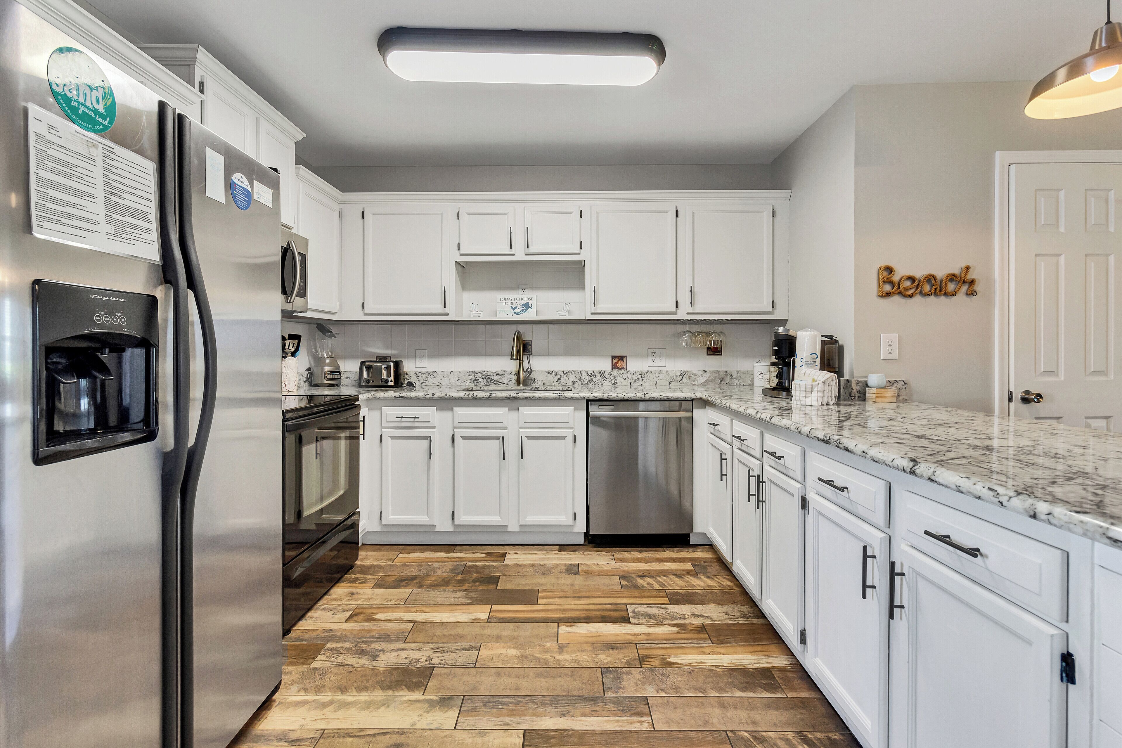 A modern kitchen with white cabinets, a stainless steel refrigerator, a dishwasher, and a marble countertop.