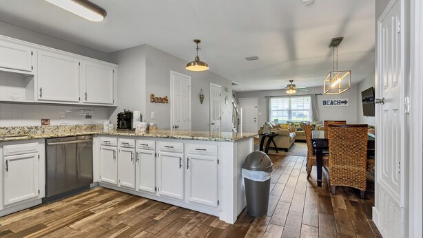 A modern kitchen with white cabinets, a granite countertop, and a wooden floor. There is a trash can and a wicker chair in the dining area. - Emerald Escapes unit 41 (Miramar Beach)