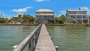 A wooden pier leading to a large, multi-story house with a balcony, surrounded by water and palm trees. - 🌅 Let's Do Sunset - Dock, Gulf Access, Sunset Deck + Pet Friendly 🐾 (Pensacola Beach)