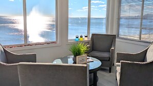 Une pièce lumineuse, baignée de soleil, avec vue sur l’océan, comprenant un ventilateur de plafond, une table ronde en verre agrémentée d’une plante, et deux fauteuils gris.