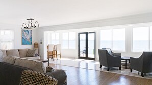 Un salon spacieux avec un grand canapé, une table basse et deux fauteuils. Il comprend également un coin repas avec une table et des chaises, et offre une vue sur l’océan grâce à de grandes fenêtres.