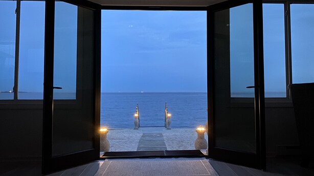 Une porte ouverte donne sur une plage, d’où l’on peut admirer la mer et un ciel dégagé.