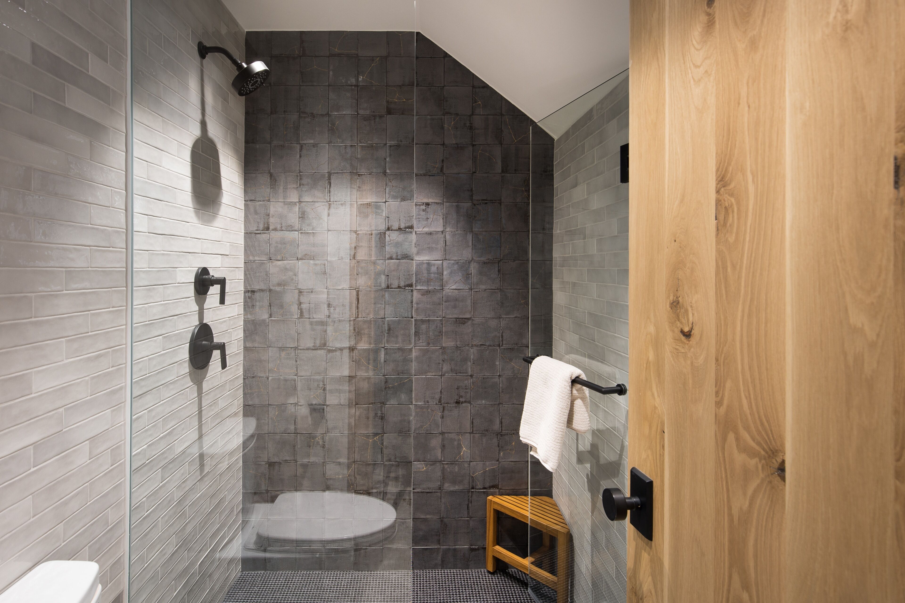 A modern bathroom with a glass-enclosed shower, a wooden door, and a small wooden stool.