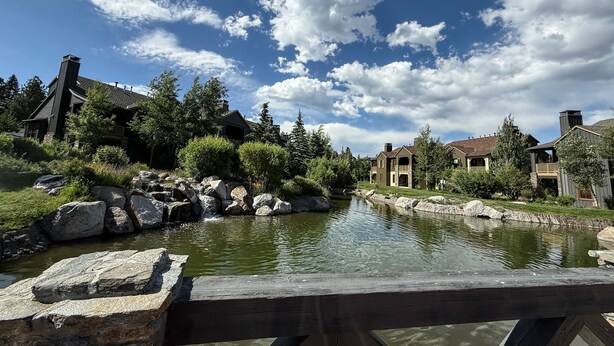 A riverside view with a wooden bridge, stone rocks, and buildings with a rustic architectural style. - Snowcreek VI The Lodges 1127 3 Suites, Spacious with Mountain Views, Private Garage and Washer Dryer by RedAwning (Mammoth Lakes)