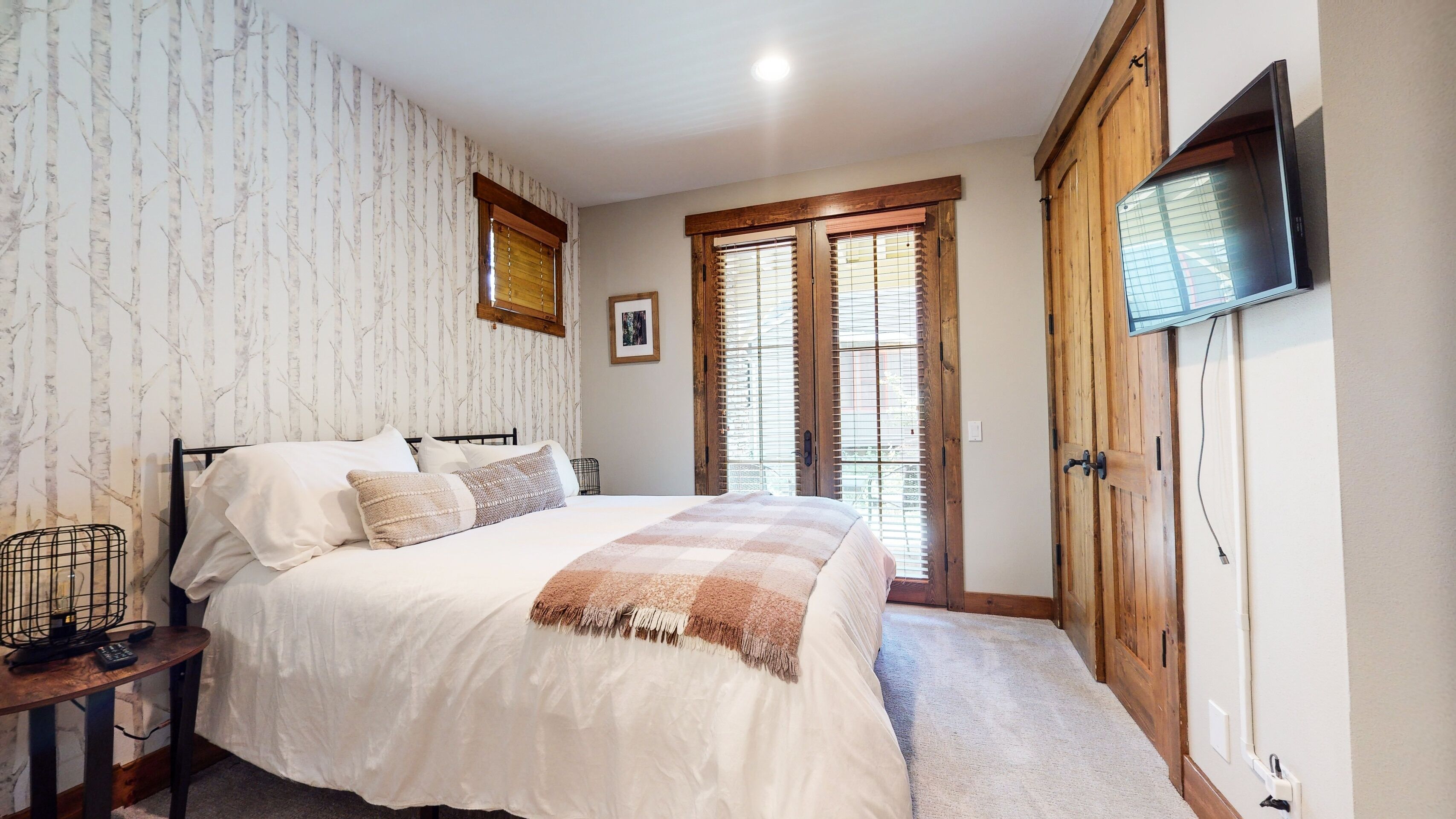 A bedroom with a bed, a wooden wardrobe, a television, and a window with blinds.