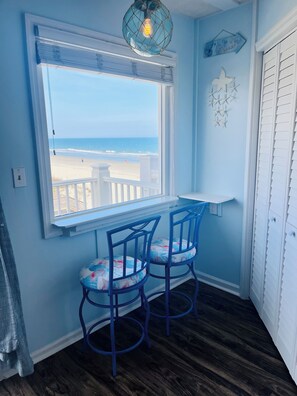 A room with a window overlooking a beach, two blue chairs, a white shelf, and a decorative wall piece.