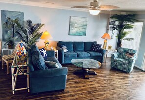 A living room with a blue sofa, a coffee table, and a chair with a nautical theme. There are two lamps, a painting on the wall, and several potted plants.