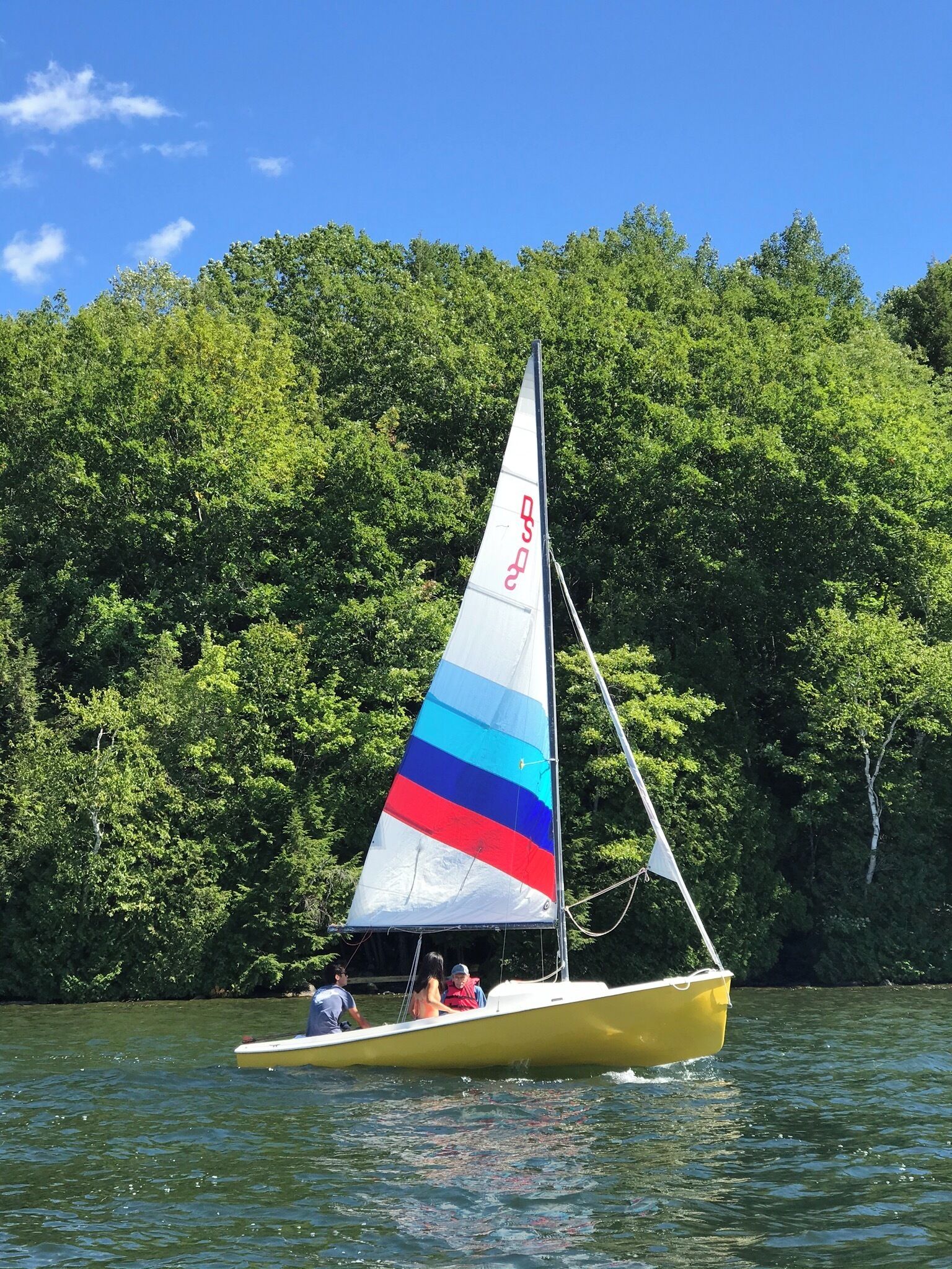 A yellow sailboat with a colorful sail on a lake surrounded by dense green trees.