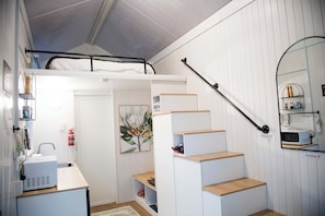 A compact room with a bunk bed, a desk with a computer, a microwave, and a staircase.